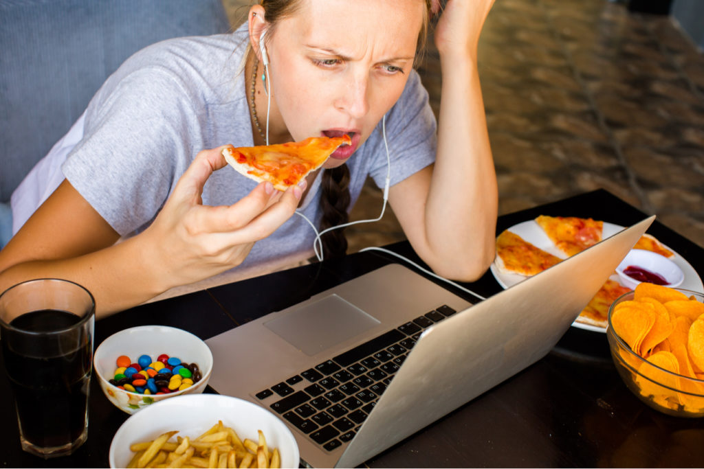 young woman eating pizza, french fries, candy, and soda while on computer wearing headphones